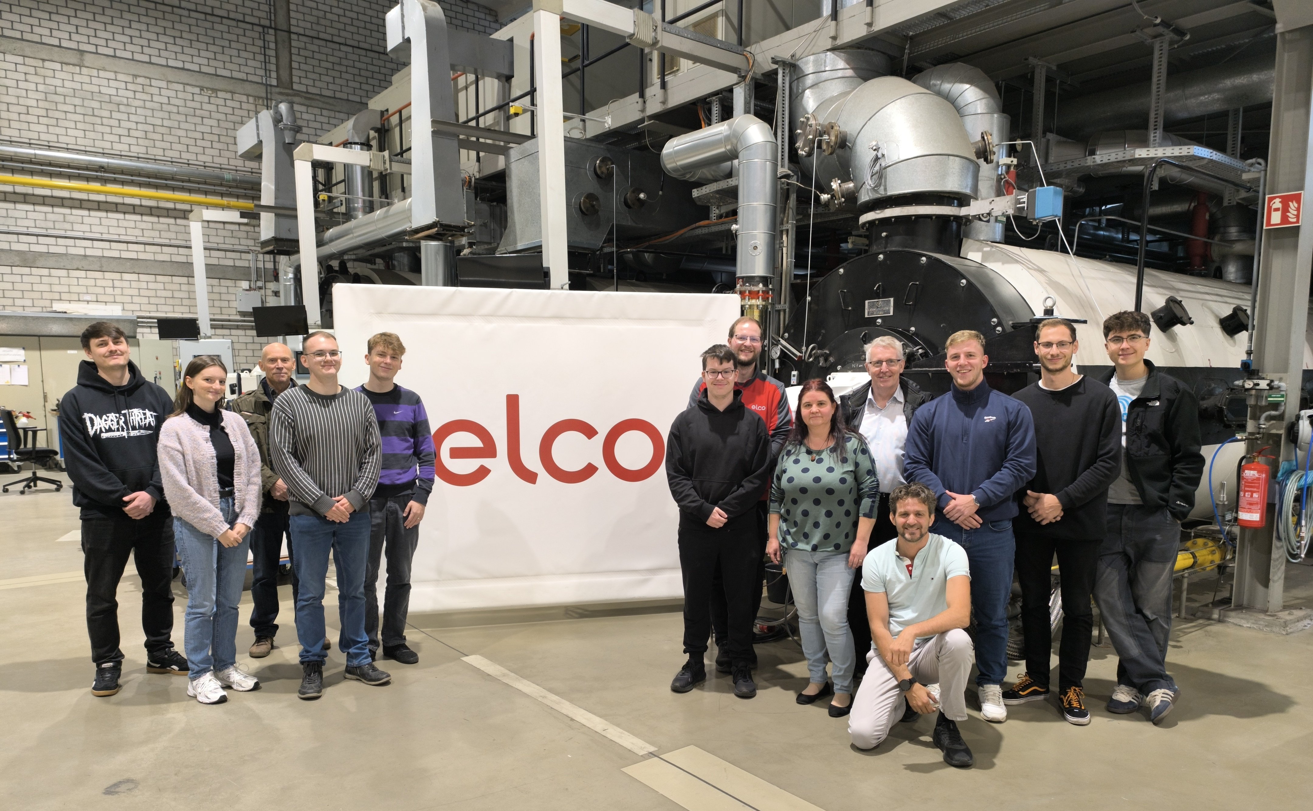 Gruppe von Menschen in einer Industriehalle vor einer großen Maschine und einem Banner mit rotem Schriftzug 'elco'.