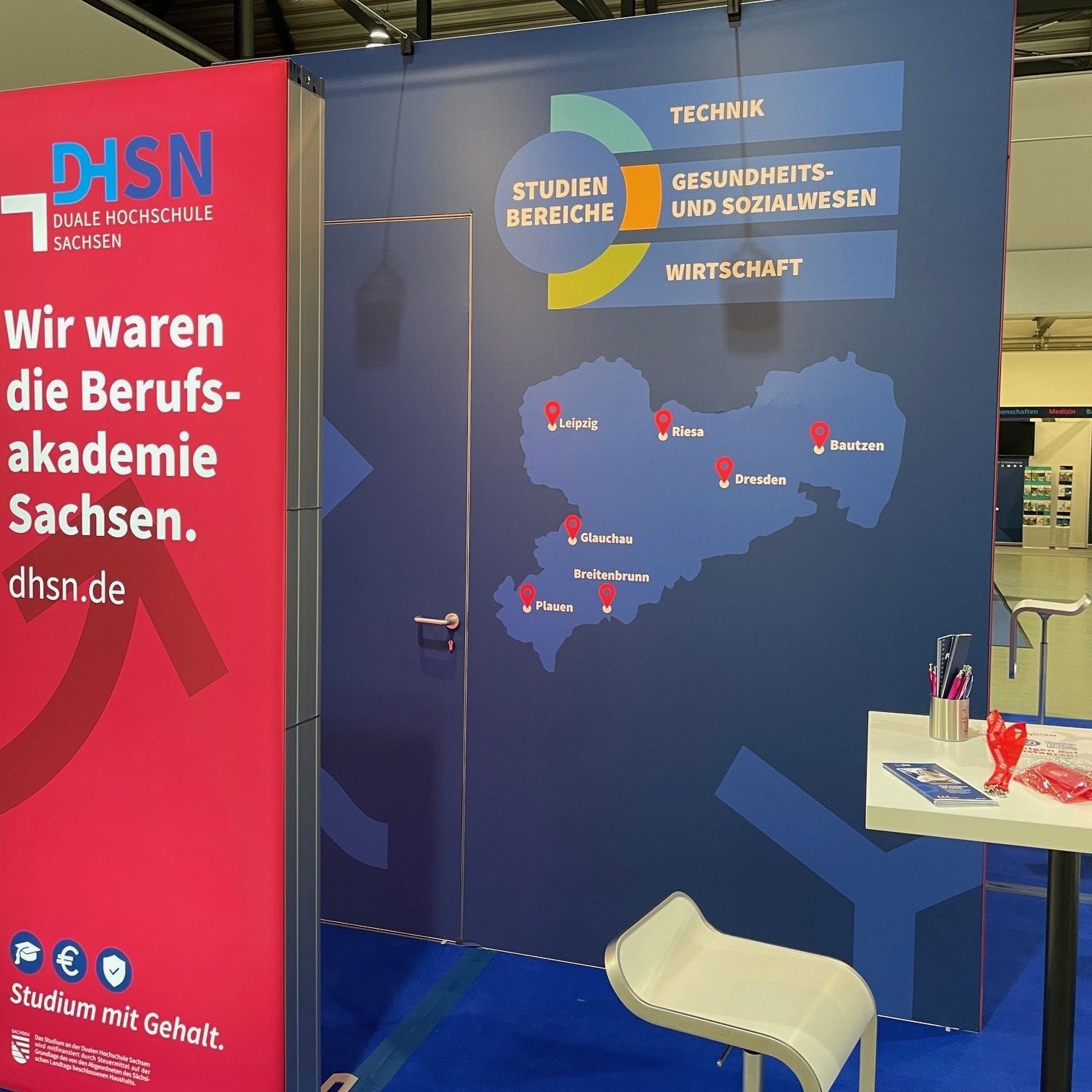 Es ist der blau rote Messestand der DHSN. Vor der Messewand steht ein Stuhl sowie ein Tisch. Auf dem Tisch liegen Werbeartikel