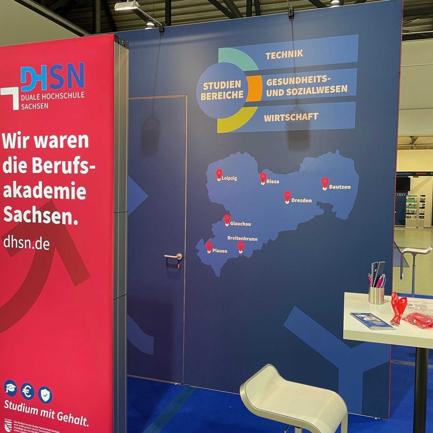 Es ist der blau rote Messestand der DHSN. Vor der Messewand steht ein Stuhl sowie ein Tisch. Auf dem Tisch liegen Werbeartikel