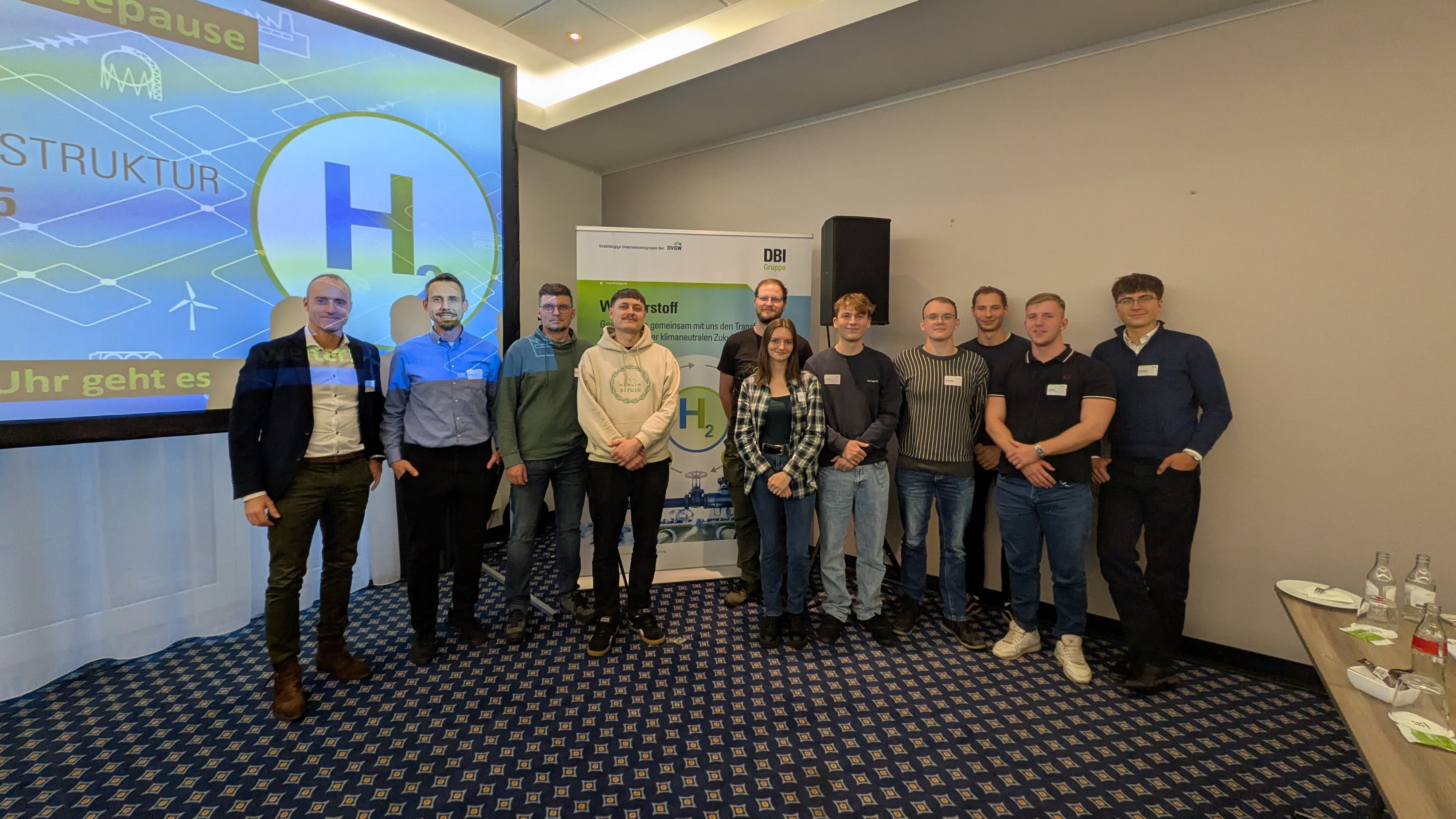 Gruppe von elf Personen steht in einem Konferenzraum vor einem Bildschirm und einem Banner mit dem DBI-Logo und dem Symbol für Wasserstoff (H2).