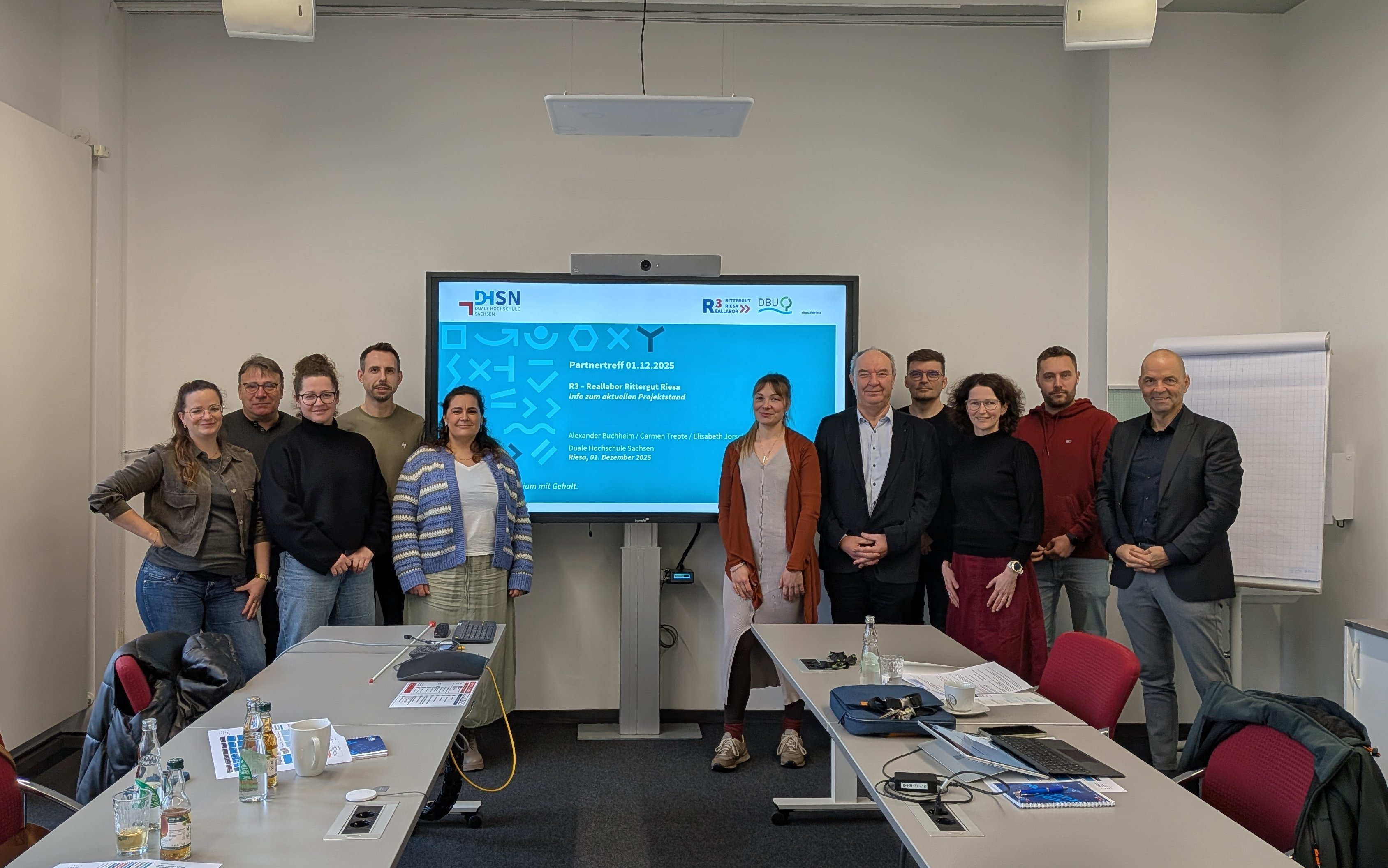 Gruppe von elf Personen steht in einem Besprechungsraum vor einem Bildschirm mit Präsentation