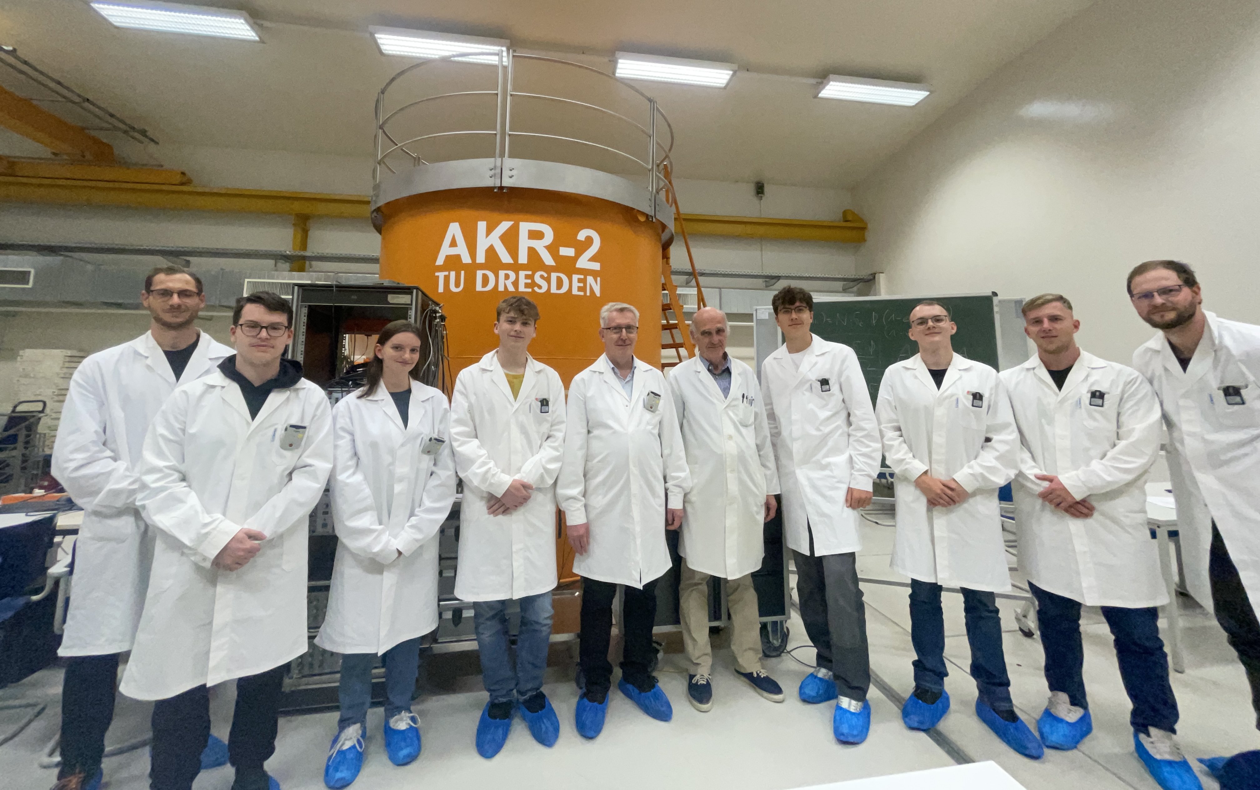 Gruppe von neun Personen in weißen Laborkitteln und blauen Überschuhen vor orangefarbenem Behälter mit Aufschrift 'AKR-2 TU DRESDEN'.