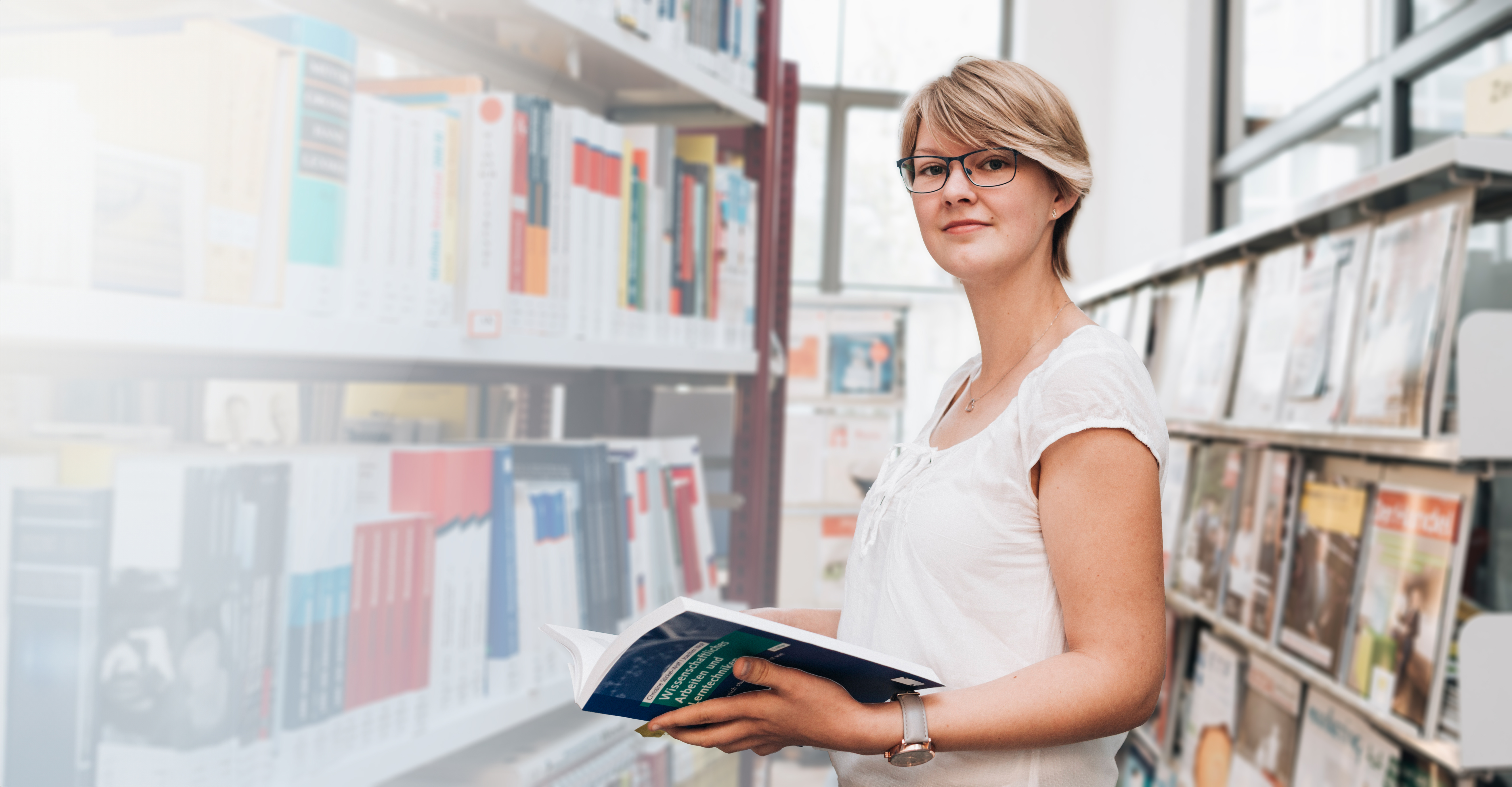Junge Frau mit Brille hält ein aufgeschlagenes Buch in einer Bibliothek vor einem Regal mit Fachliteratur.