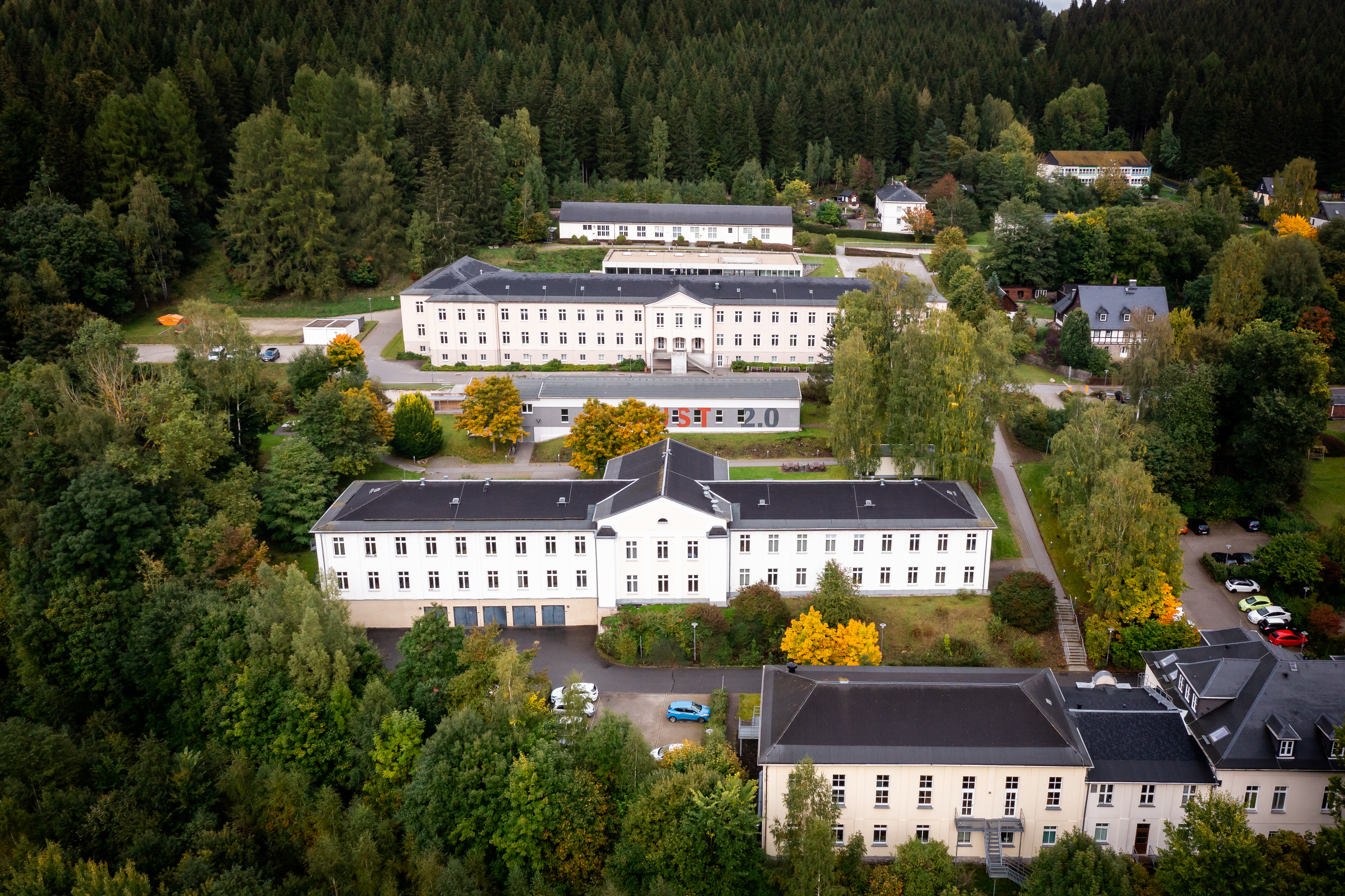 Luftaufnahme einer weitläufigen, weißen Gebäudeanlage, umgeben von dichtem Wald. Mehrere längliche Gebäude mit dunklen Dächern und vielen Fenstern sind in einer Reihe angeordnet. Einige Bäume mit gelbem Herbstlaub sind verteilt. Parkplätze vor den Gebäuden. Im Vordergrund und Hintergrund dichter, grüner Wald. (automatisch generiert)