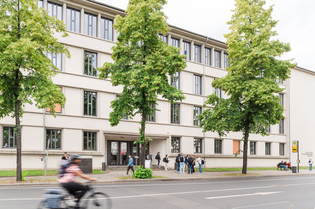 Auf dem Bild sieht man ein Gebäude. Das Gebäude ist der Standort Dresden der Dualen Hochschule Sachsen und im Vordergrund ist eine Radfahrerin verschwommen zu erkennen.