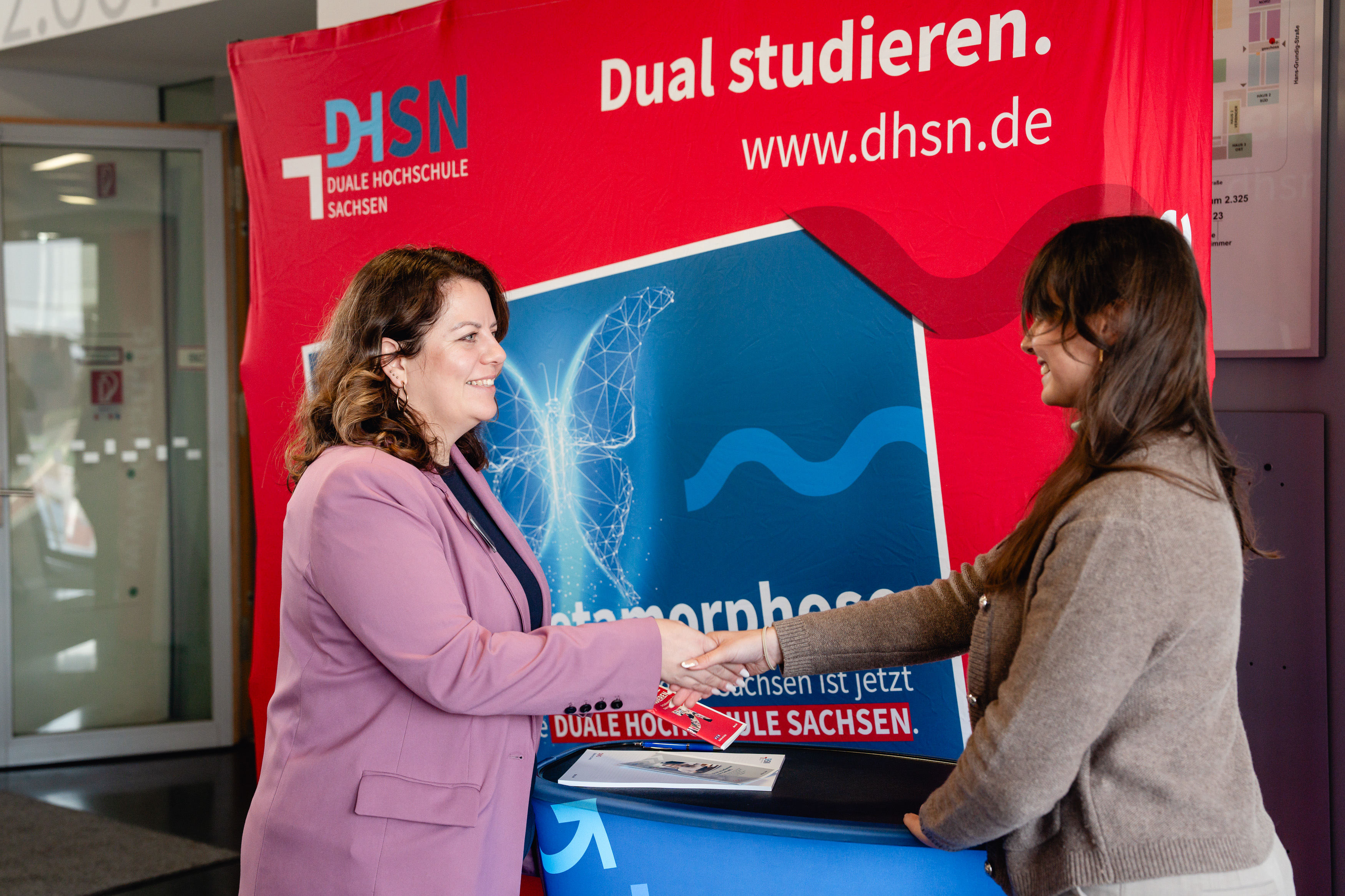  Es ist ein Messestand der Dualen Hochschule zu sehen. Im Vordergrund sind eine Schülerin zu sehen, die mit einer Beraterin am Stand spricht. Im Hintergrund ist die rote Messewand mit weißer Schrift zu sehen. 