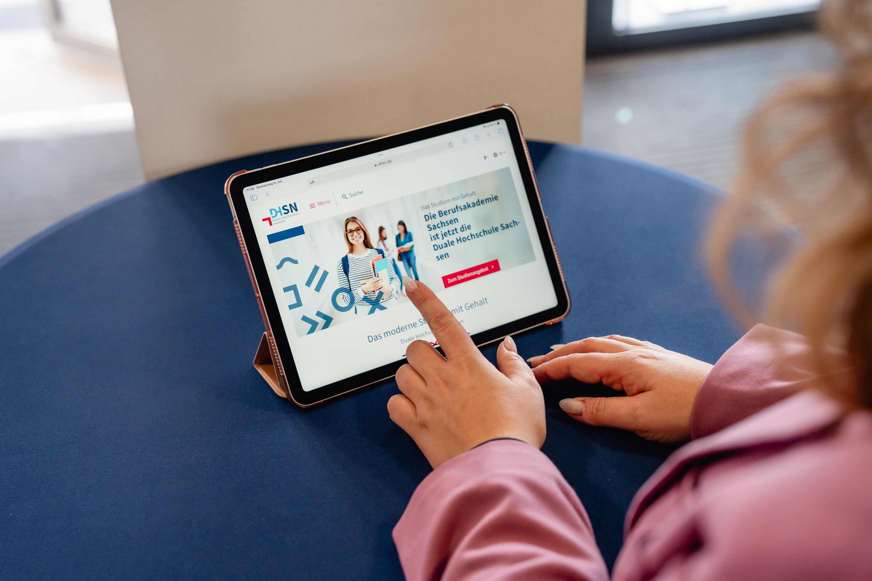 Eine Frau tippt auf ein Tablet. Das Tablet steht auf einem blauen Tisch. Auf dem Tablet ist die Webseite der DHSN zu sehen