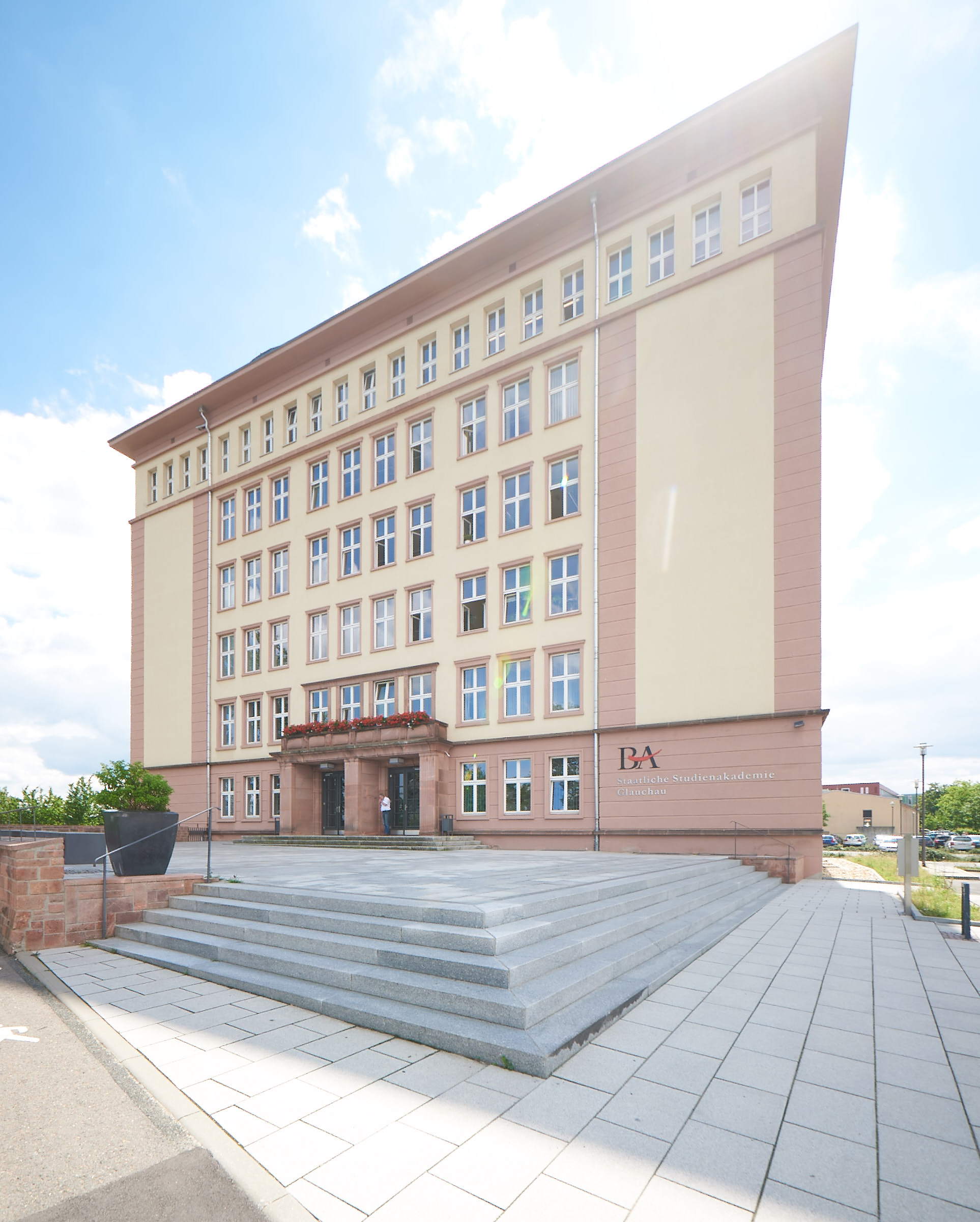 Dreigeschossiges Gebäude mit roter Klinkerfassade und hellgelben Putzstreifen zwischen den Fenstern. Vor dem Gebäude breite, hellgraue Stufenanlage mit Podest und einer Person vor der Eingangstür. Auf dem obersten Podest ein Schild mit der Aufschrift BA. Links im Bild ein kleiner Baum in einem Pflanzgefäß. Rechter Bildrand geparkte Autos. Heller Himmel. (automatisch generiert)