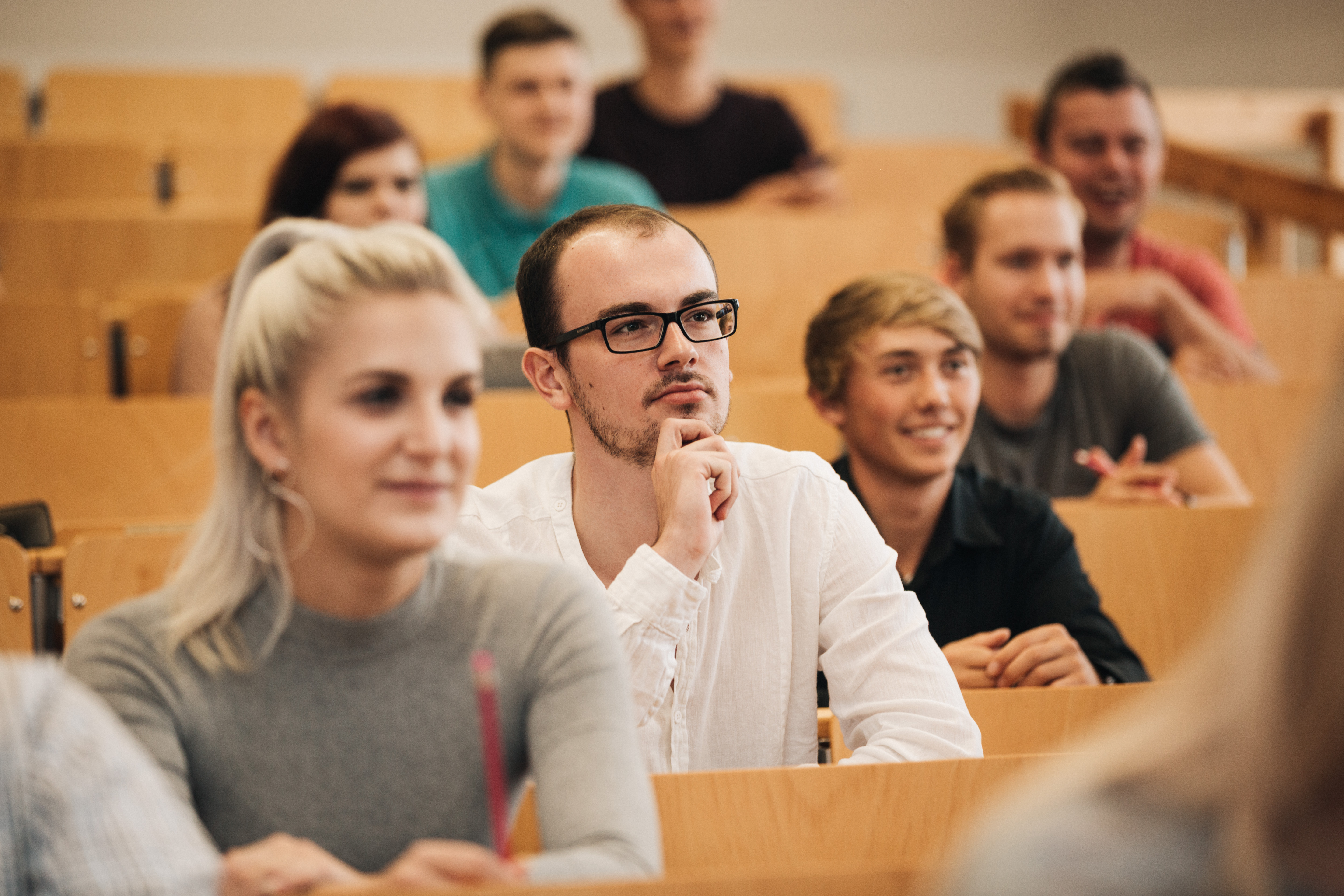 Reihe von jungen Erwachsenen in einem Hörsaal, der Mann im Vordergrund mit Brille und weißem Hemd blickt nachdenklich nach oben, die anderen lächeln oder zeigen Interesse. (automatisch generiert)