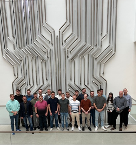 Eine Gruppe von etwa 20 jungen Männern und wenigen jungen Frauen steht vor einem großen, weißen Wandrelief mit einem abstrakten, geometrischen Muster aus vertikalen Linien. Die meisten Personen tragen Jeans und dunkle Oberteile. Der Hintergrund ist eine weiße Wand und eine Treppe links im Bild. (automatisch generiert)