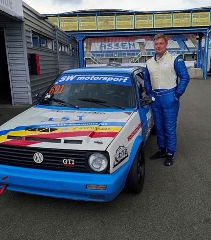 Rennwagen VW Golf GTI, blau-weiß kariert, mit Rennaufklebern, steht auf Asphalt. Ein Mann in Rennkleidung lehnt lächelnd davor. Im Hintergrund eine Tribüne und Industriegebäude. (automatisch generiert)