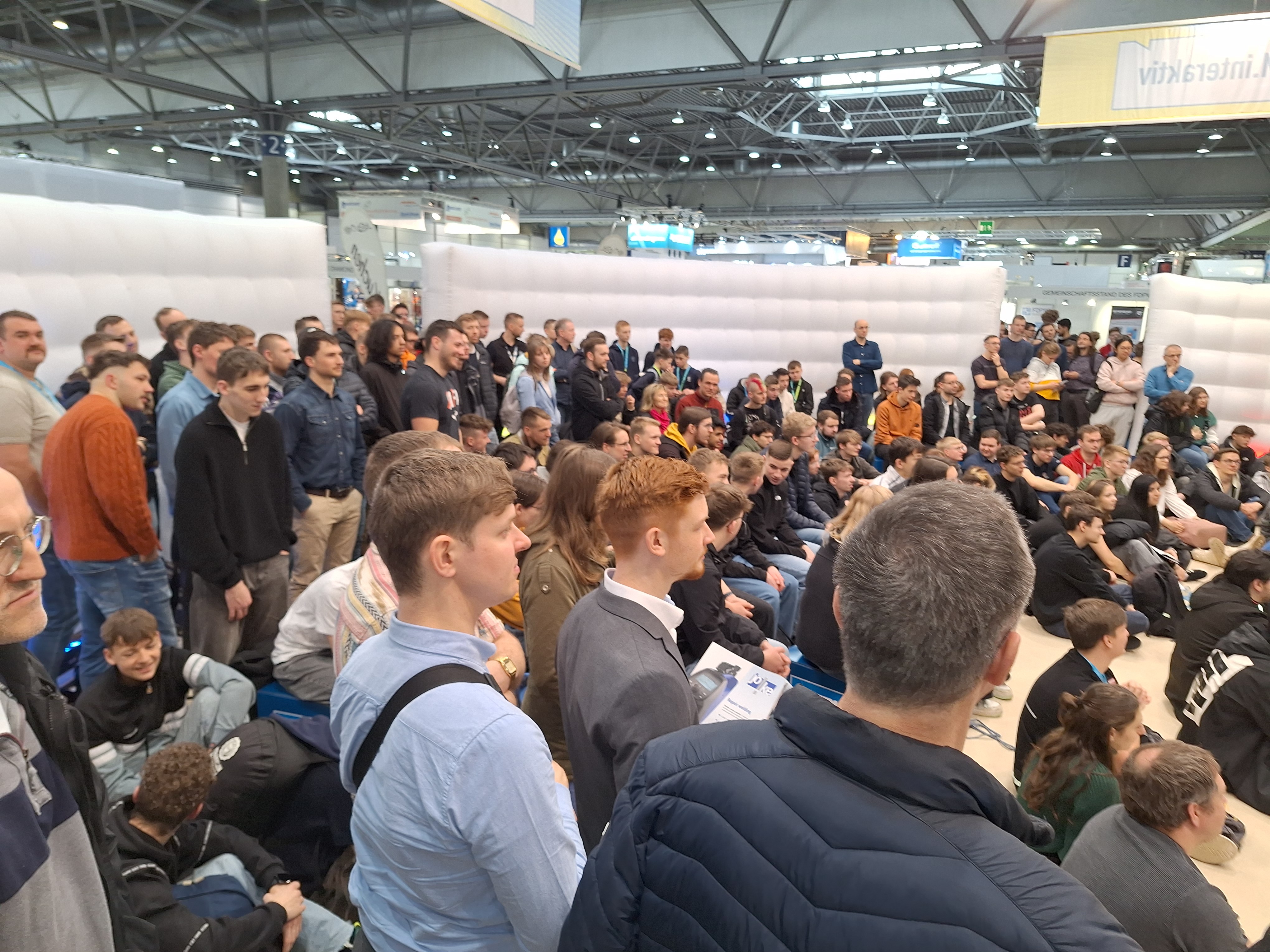 Große Menschengruppe sitzt auf dem Boden und steht vor einer Bühne in einer hellen, industriell wirkenden Halle. Die Mehrheit sind junge Männer, viele tragen Anzug oder Hemd. Einige halten Notizen oder schauen auf ihre Handys. Im Hintergrund eine Bühne und Banner. Die Beleuchtung ist hell, das Licht scheint von der Decke. (automatisch generiert)