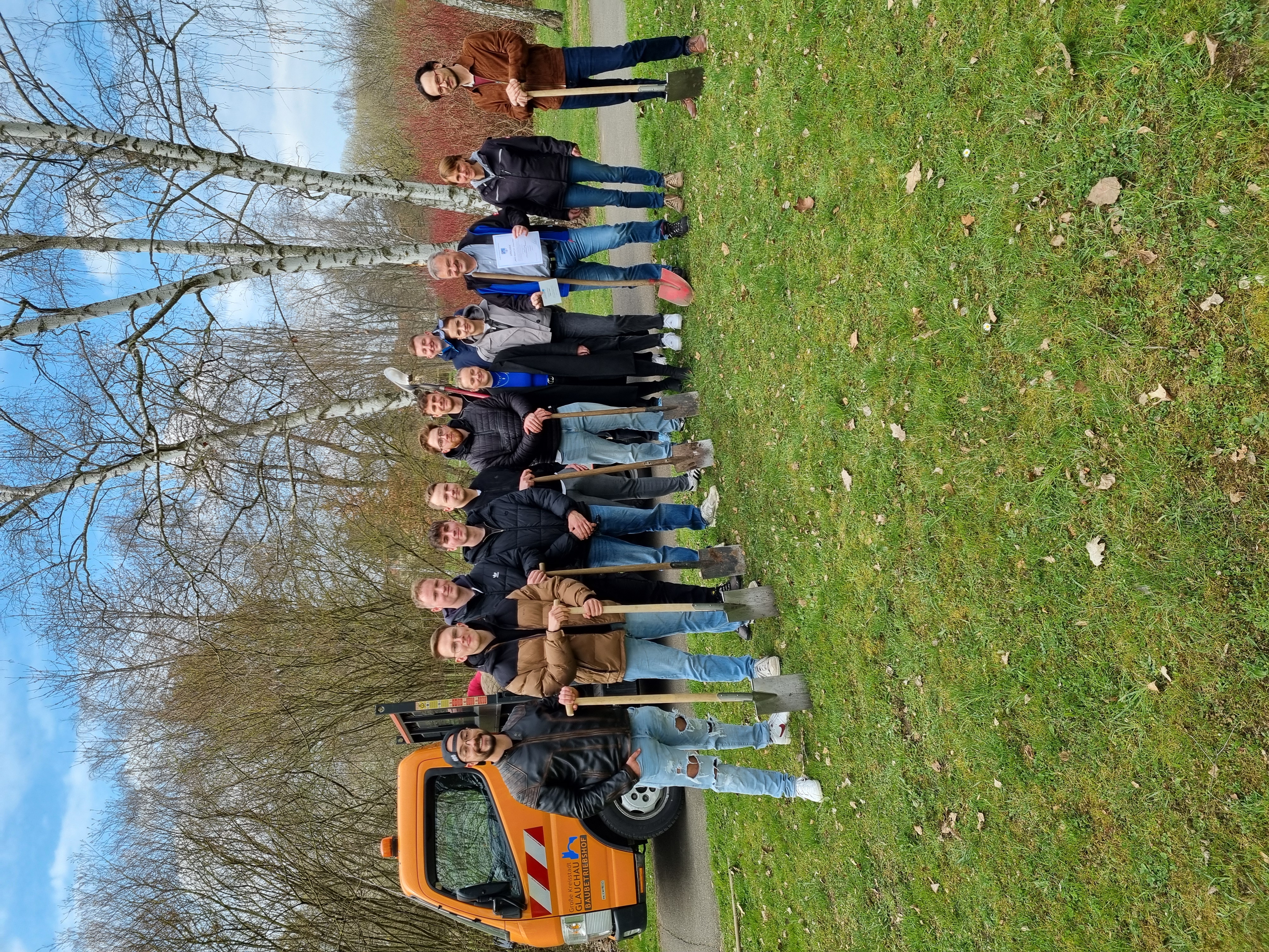 Eine Gruppe von etwa zwanzig Männern steht in einer Reihe vor einem orangefarbenen Gerätewagen. Alle tragen dunkle Arbeitskleidung und halten jeweils einen langen, dünnen Stab in der Hand. Der Wagen ist mit dem Schriftzug "FORST BAUER" versehen. Im Hintergrund sind Bäume und ein blauer Himmel zu sehen. Die Aufnahme wurde aus Vogelperspektive gemacht. (automatisch generiert)