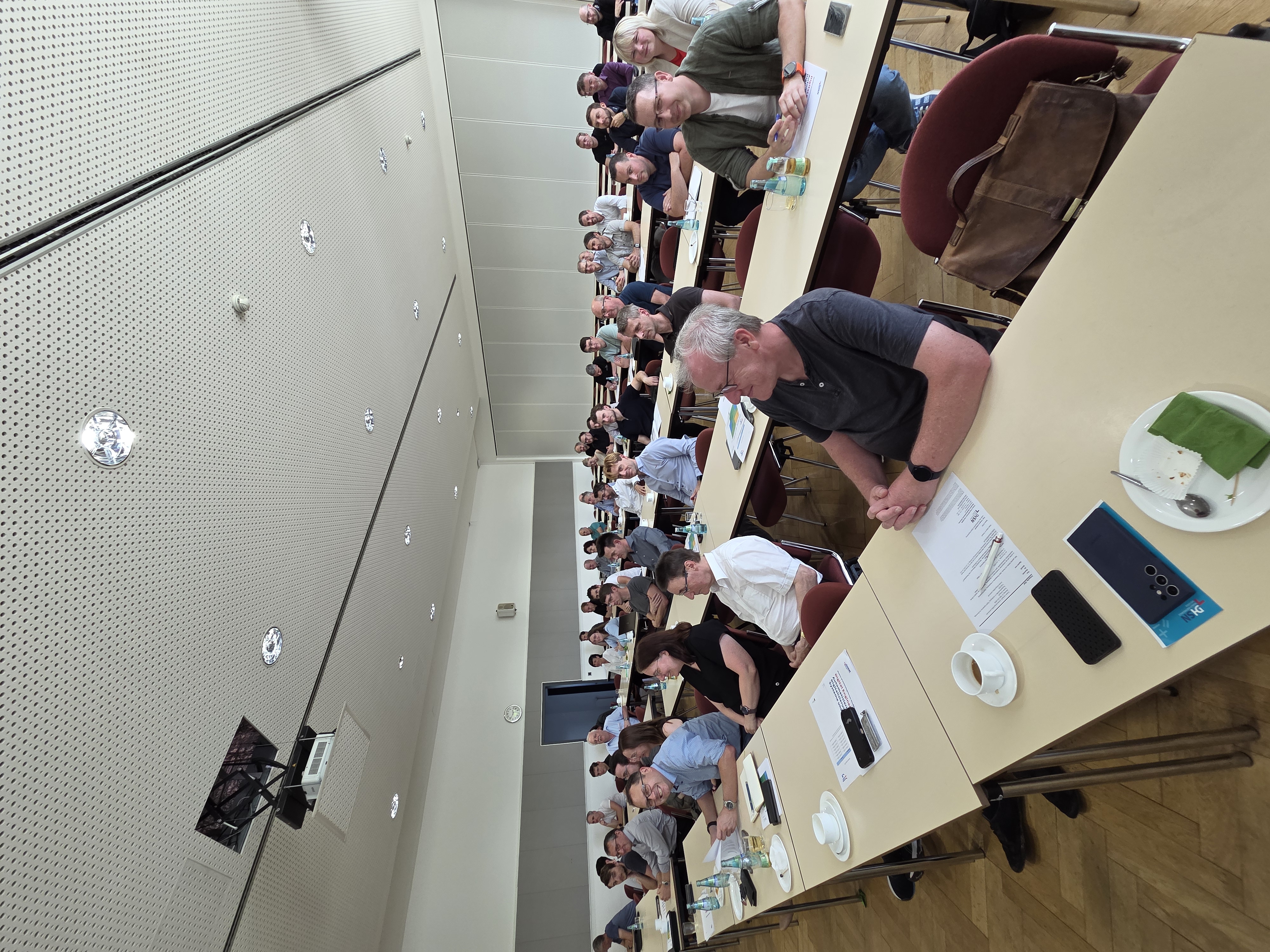 Draufsicht auf eine lange Reihe von Personen, die an Tischen in einem Hörsaal sitzen. Die Personen sind hauptsächlich Männer mittleren Alters, die an einer Konferenz oder einem Seminar teilnehmen. Auf den Tischen befinden sich Notizblöcke, Stifte, Wasserflaschen und elektronische Geräte. Im Vordergrund ein Mann in schwarzem T-Shirt, der etwas auf einem Tablet liest. Im Hintergrund sind mehrere Reihen von Tischen und Stühlen zu sehen. Der Hörsaal hat eine gepunktete Wand und Decke und ist mit mehreren Lichtquellen ausgestattet. (automatisch generiert)