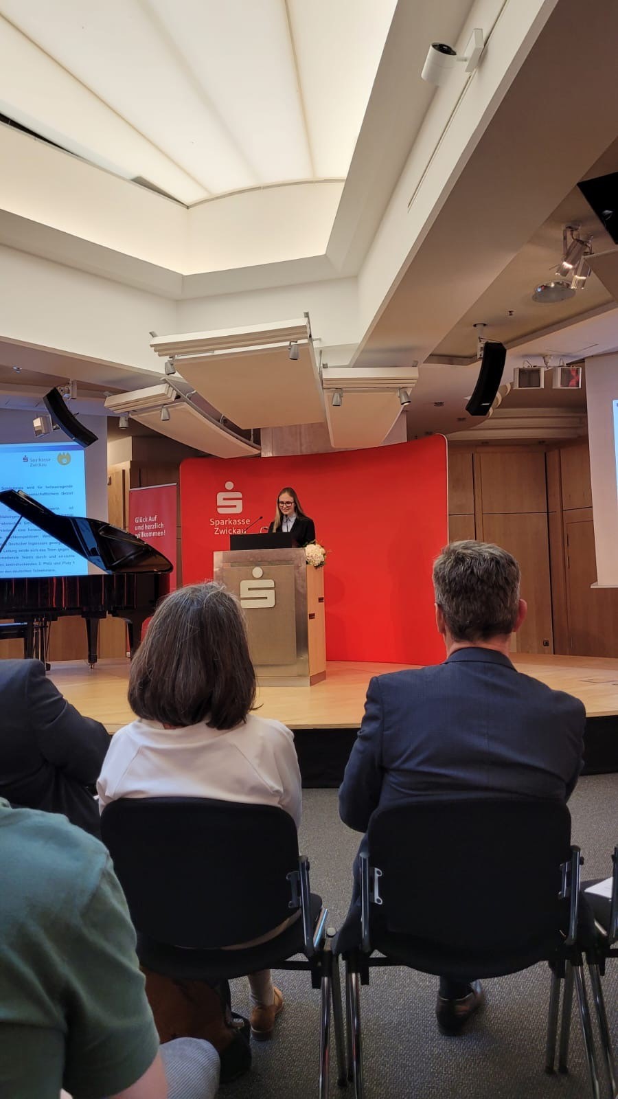 Innenraum eines Konzertsals mit rotem Podium. Eine Person steht am Rednerpult und spricht. Vor dem Podium sitzen mehrere Personen auf schwarzen Stühlen und schauen zum Redner. Im Hintergrund ein Flügel und Werbebanner der Sparkasse. Lichtquellen sind in der Decke installiert. (automatisch generiert)