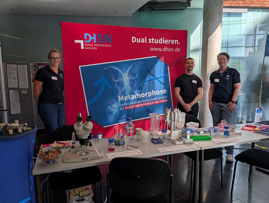 Der Stand der Staatlichen Studienakademie zur Langen Nacht der Wissenschaften in der BIO CITY Leipzig. Im Bild sind drei Personen zu sehen. Von rechts nach links: Laboringenieurin Melanie Thielemann, Biotechnologie-Studierender Kurt Mühlberg und Dozentin Barbe Rentsch. Im Vordergrund befinden sich Laborgeräte, ein Mikroskop und weitere typische Laborutensilien.