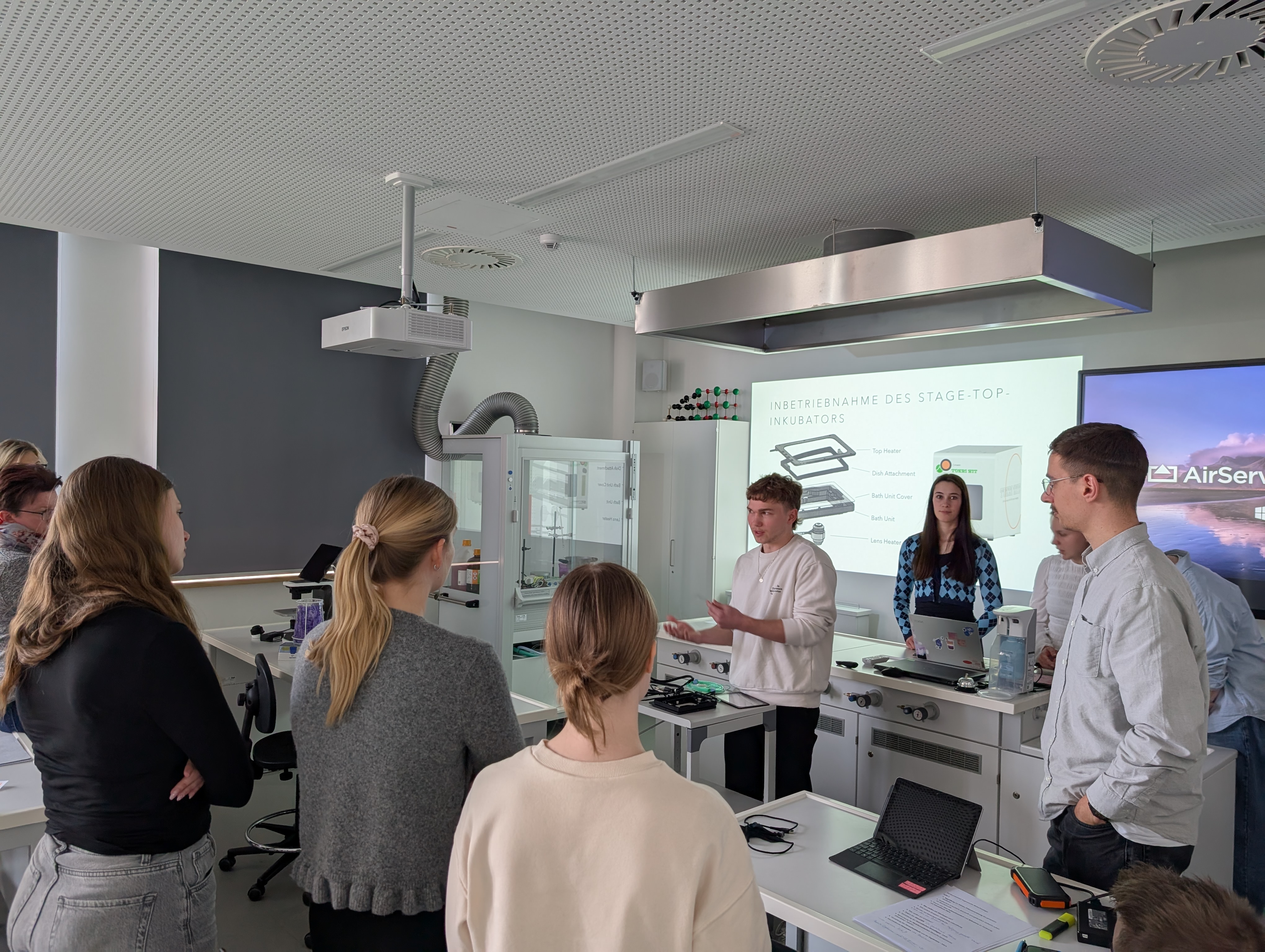 Das Bild zeigt Studierende des Studiengangs Labor- und Verfahrenstechnik, Fachrichtung Biotechnologie, in einem modernen Labor. Ein junger Mann präsentiert vor Kommilitonen Forschungsergebnisse. Eine Projektion zeigt die 'INBETRIEBNAHME DES STAGE-TOP-INKUBATORS' für die Live-Zellanalyse. Die Szene verdeutlicht die praxisorientierte und forschungsnahe Lernumgebung, in der im 6. Semester reale Laborprojekte umgesetzt werden.