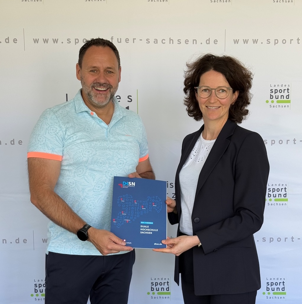 Links steht Christian Dahms in hellblauem Poloshirt und rechts daneben steht Frau Prof. Dr. Katja Soyez in dunklem Anzug, sie halten gemeinsam ein blaue Mappe der DHSN in den Händen und stehen vor einer Wand mit Logos des Landessportbunds Sachsen.