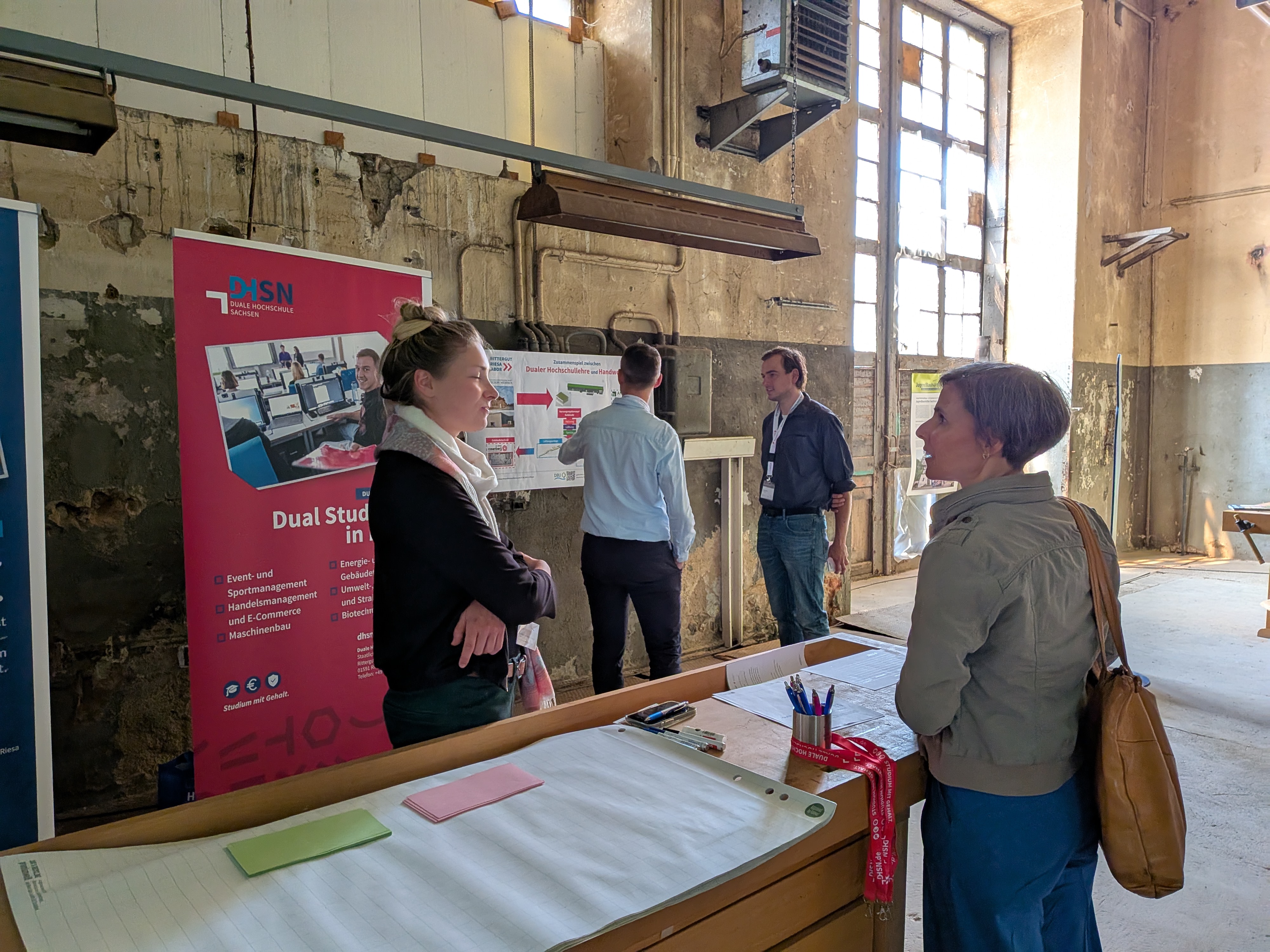 Mehrere Personen stehen vor einer Werkbank und betrachten eine Wand mit Plakaten in einem hellen Raum mit großen Fenstern.