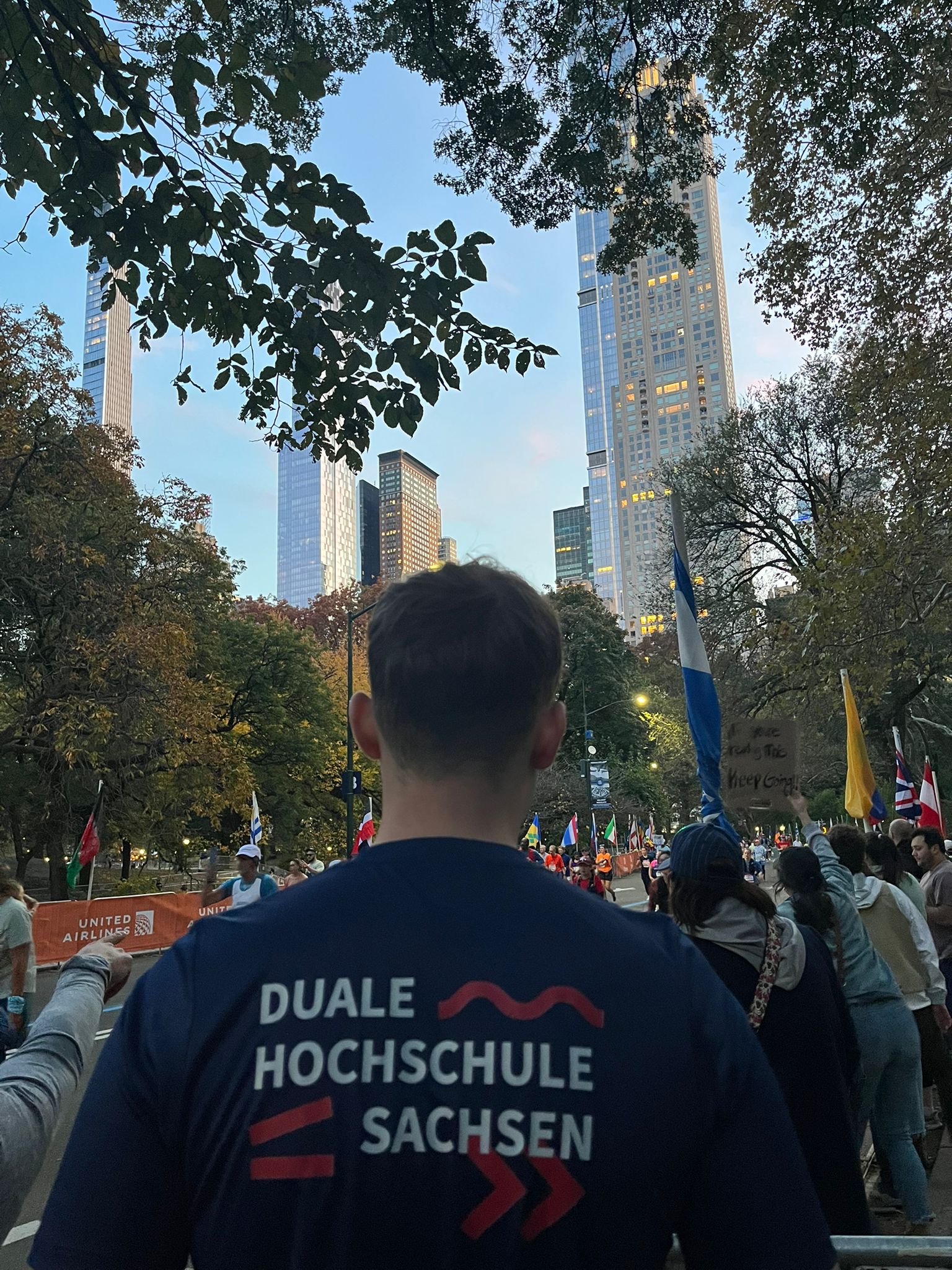 Robin Trojan steht im DHSN-Shirt am Zieleinlauf des Marathons im Central Park mit den Läufern, Läuferinnen und New Yorker Wolkenkratzern im Hintergrund.